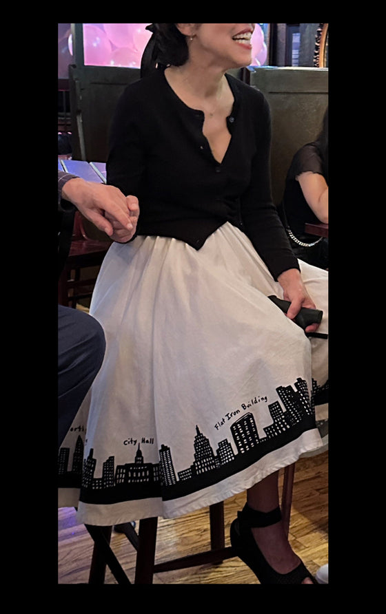 Person wearing a black and white dress with a city skyline design, sitting in a bar.