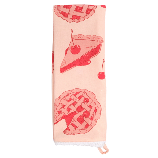 Pale pink tea towel folded lengthwise and hanging vertically, decorated with red illustrations of cherry pie slices, lattice-top pies, and cherries, with white lace trim at the bottom.