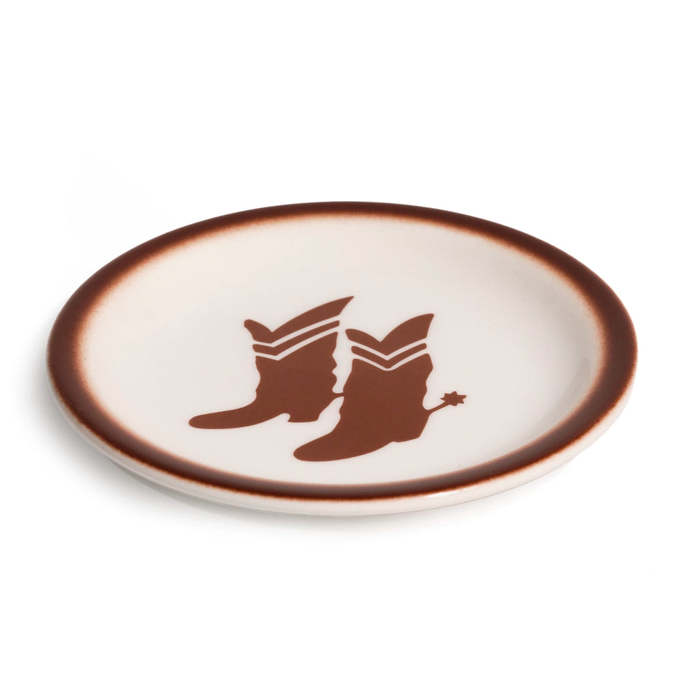 Angled view of a cream glazed ceramic plate with a brown cowboy boots-and-spur graphic and shaded brown rim, showing the gently raised edge and roomy serving surface.