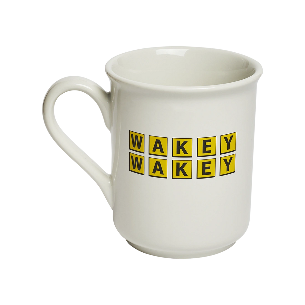 Classic white ceramic mug with “WAKEY WAKEY” printed in bright yellow tile-style lettering. Playful coffee mug perfect for morning routines, caffeine enthusiasts, and breakfast tables. Angled view.