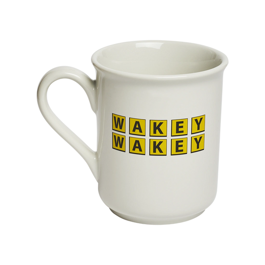 Classic white ceramic mug with “WAKEY WAKEY” printed in bright yellow tile-style lettering. Playful coffee mug perfect for morning routines, caffeine enthusiasts, and breakfast tables. Angled view.