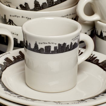 White mug with black city skyline design on a matching saucer, surrounded by other similar mugs.