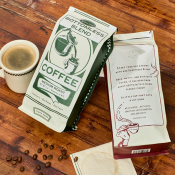 Two coffee packages on a wooden surface with a cup of coffee and coffee beans.