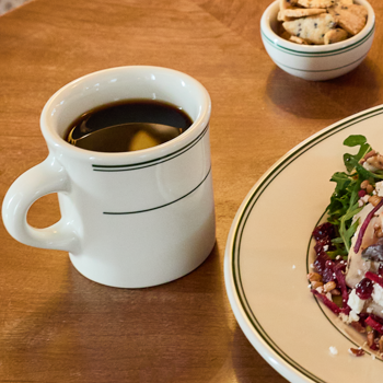 Mug of coffee on a wooden table with a plate of food and a bowl of crackers.