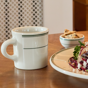White mug with green rim on a wooden table next to a plate of food and a small bowl.