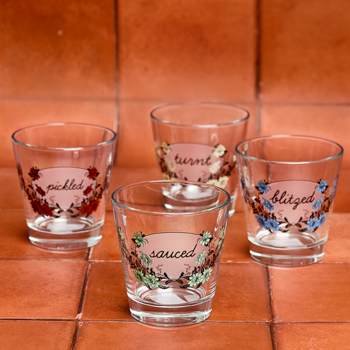 Set of four glass tumblers with printed text and floral designs on a terracotta tile surface.