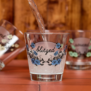 Glass with floral design and 'blitzed' text being filled with a liquid on a wooden surface.