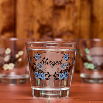 Clear shot glass with floral design and 'blitzed' text on a wooden surface