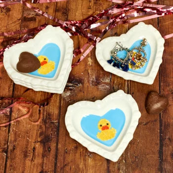 Three heart-shaped duck dishes holding chocolate and earrings on a table with pink ribbon