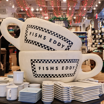 Stack of white ceramic mugs with 'Fishes Eddy' branding in a store setting.