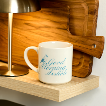 Mug with 'Good Morning Asshole' text on a wooden shelf with a lamp and cutting board.