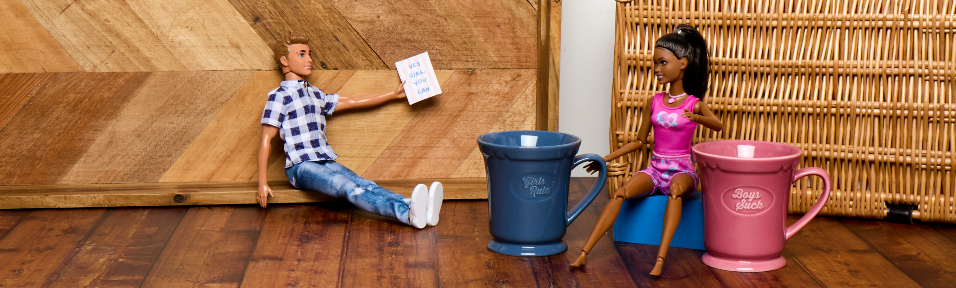 Two dolls, one male and one female, sitting on a wooden floor with two large mugs in front of them.