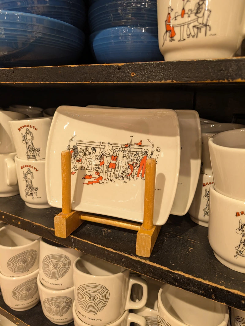 Liana Finck subway ride tray on a wooden stand among cream mugs