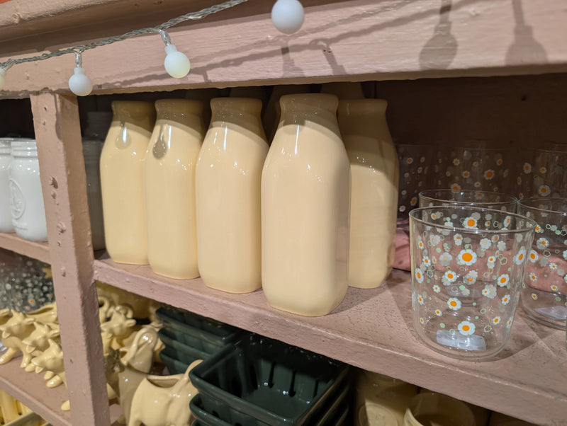 Row of butter yellow milk jugs on a pink shelf beside a daisy glass