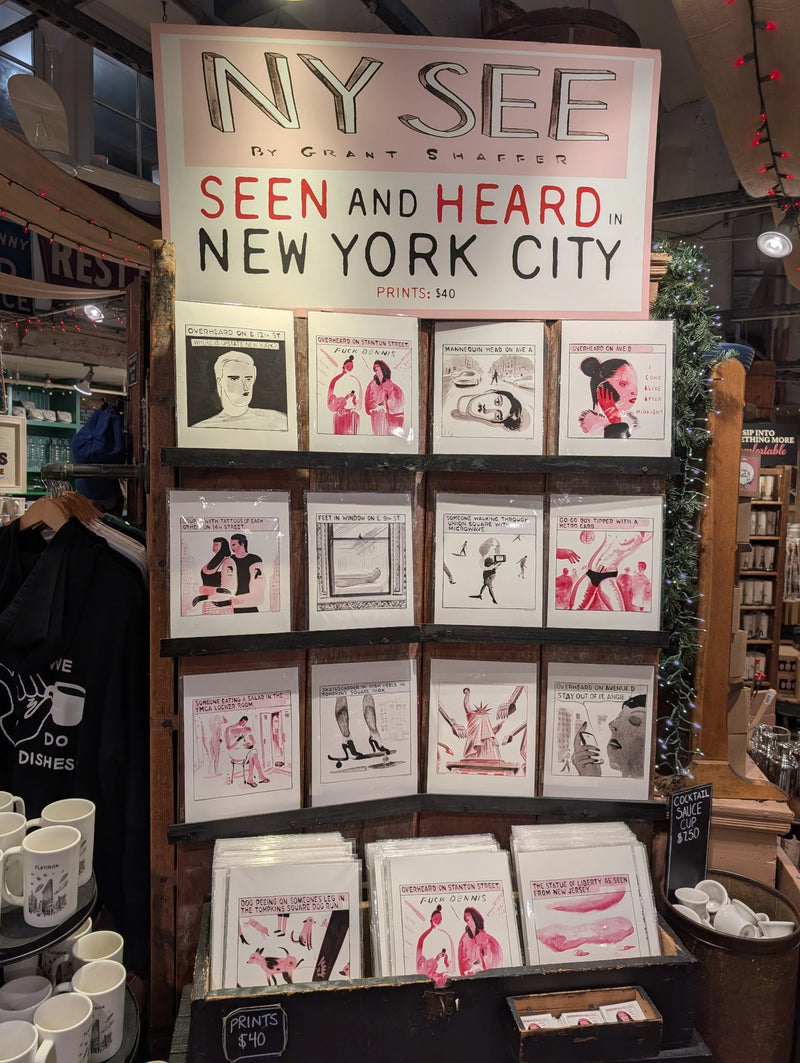 Wall display of NYC art prints under a NY SEE sign, with Flatiron mugs below
