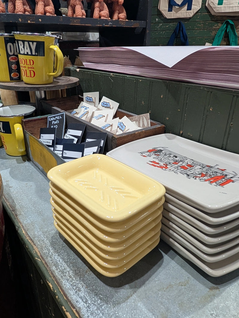 Stack of butter yellow soap dishes on a store counter beside printed trays